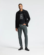 A man exudes confidence as he poses against a plain backdrop, clad in a Psycho Bunny Men's Dumon Graphic Tee in black, made from premium Pima cotton. He sports a black jacket over it, paired with greenish-gray pants and black boots. With one hand casually tucked in his pocket, he strikes a relaxed stance.