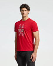 A man is wearing a red MENS DUMON GRAPHIC TEE - B6U271E200 by Psycho Bunny, showcasing a distinctive bunny head and crossbones logo. Made from high-quality Pima cotton, the shirt offers both comfort and style as he stands against a simple, light-colored background.