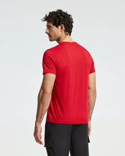A man with curly hair stands facing slightly away, wearing a vibrant red Psycho Bunny Men's Dumon Graphic Tee (B6U271E200) crafted from 100% Peruvian Pima cotton. The plain background accentuates the striking hue and premium feel of his attire.