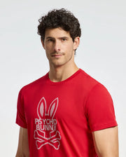 A person with curly hair wears a red Psycho Bunny MENS DUMON GRAPHIC TEE - B6U271E200, crafted from premium Pima cotton. It features a bunny design with crossbones against a plain white background.