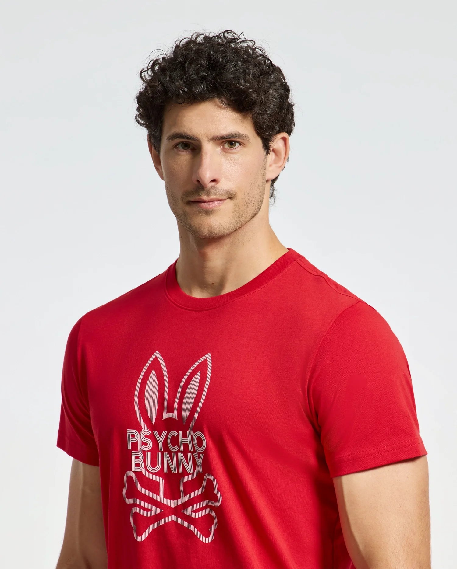 A person with curly hair wears a red Psycho Bunny MENS DUMON GRAPHIC TEE - B6U271E200, crafted from premium Pima cotton. It features a bunny design with crossbones against a plain white background.