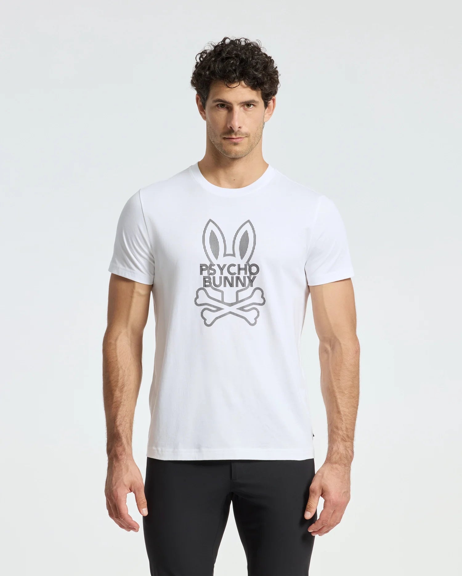 A man wears a Psycho Bunny MENS DUMON GRAPHIC TEE - B6U271E200, crafted from premium Pima cotton and adorned with the iconic Psycho Bunny logo—a bunny with crossbones. The white tee stands out against his black pants against a plain white backdrop.