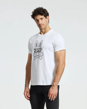 A man with curly hair is standing against a plain backdrop, wearing black pants and a white Psycho Bunny MENS DUMON GRAPHIC TEE - B6U271E200. The T-shirt is made from premium Pima cotton and showcases the iconic bunny logo complete with ears and skull and crossbones, perfectly blending style and comfort.