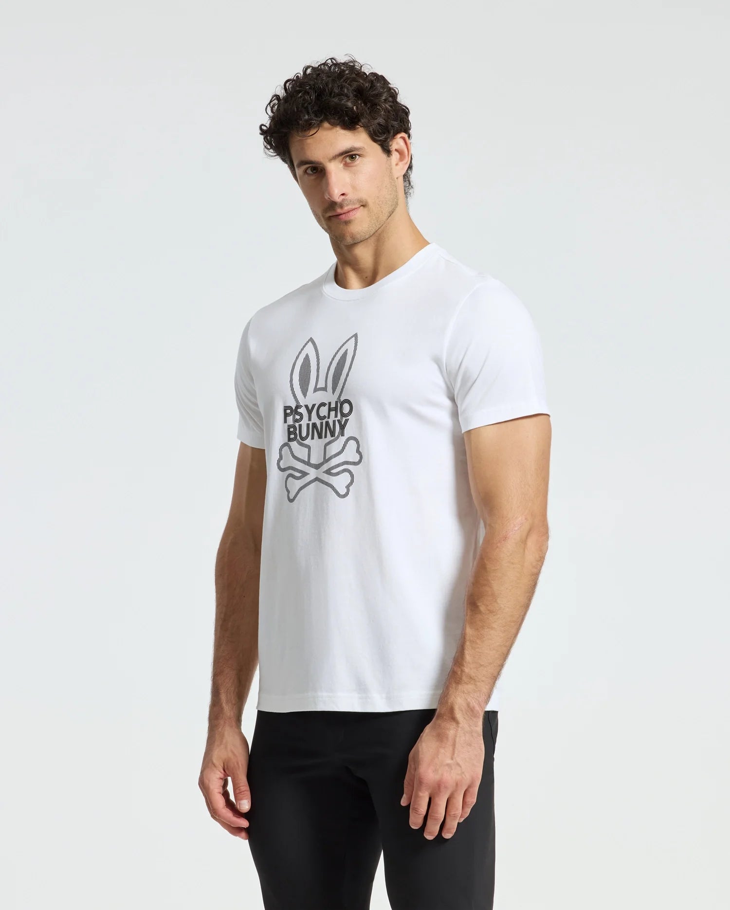 A man with curly hair is standing against a plain backdrop, wearing black pants and a white Psycho Bunny MENS DUMON GRAPHIC TEE - B6U271E200. The T-shirt is made from premium Pima cotton and showcases the iconic bunny logo complete with ears and skull and crossbones, perfectly blending style and comfort.