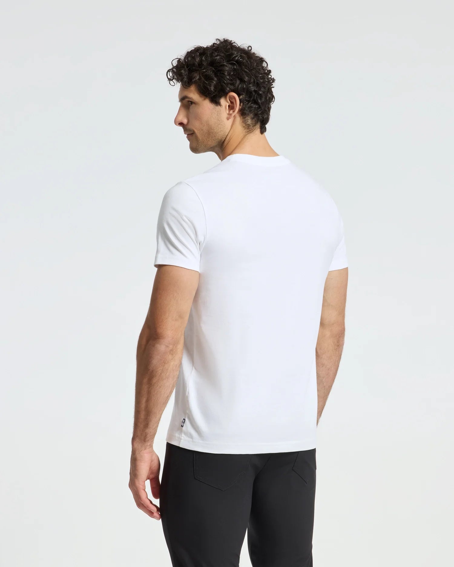 A man with curly hair stands, gazing to the side, dressed in a plain white MENS DUMON GRAPHIC TEE (Style Code: B6U271E200) from Psycho Bunny and black pants. The background is a light, neutral color.