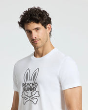 A man with curly hair is wearing a white MENS DUMON GRAPHIC TEE - B6U271E200 by Psycho Bunny, showcasing a stylized bunny logo and the words "Psycho Bunny." Crafted from Premium Pima Cotton, he looks into the camera with a neutral expression against a plain background.