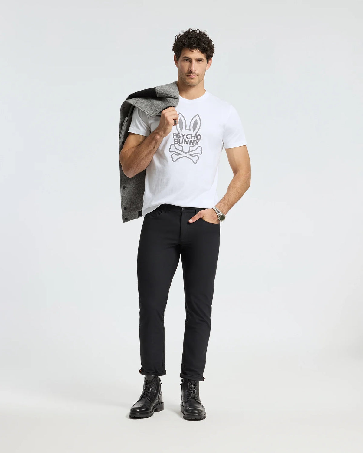 A man stands confidently against a plain background, wearing the Psycho Bunny MENS DUMON GRAPHIC TEE - B6U271E200 made from Premium Pima Cotton. He pairs it with black pants and boots, carrying a gray jacket over his shoulder. His short curly hair enhances his stylish yet casual look.