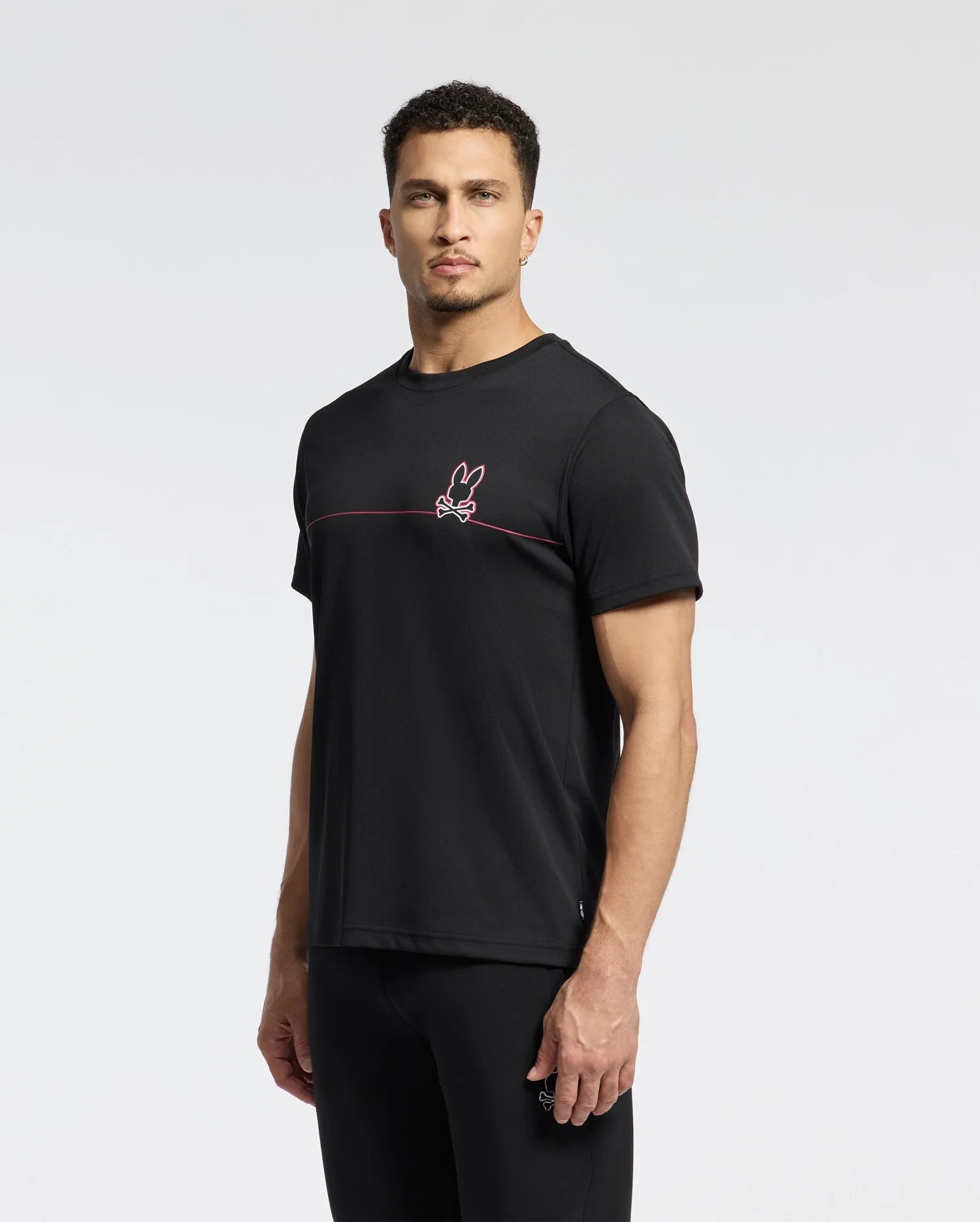A man with short curly hair poses against a white background, wearing the Psycho Bunny MENS DACANO CHEST GRAPHIC SPORT TEE - B6U313E200, featuring moisture-wicking technology, a small red and white bunny logo, and a thin red line across the chest.