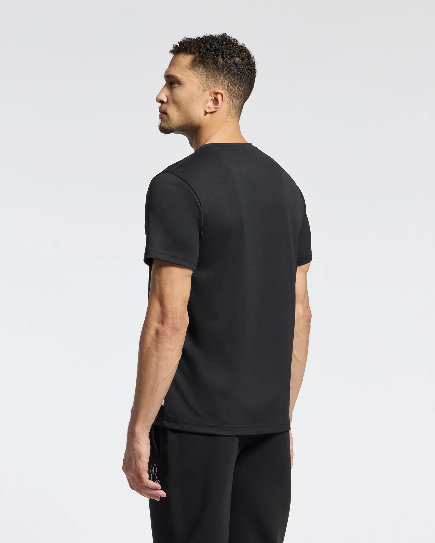 A man with short, curly hair wears the Psycho Bunny MENS DACANO CHEST GRAPHIC SPORT TEE and pants made of performance fabric. He stands against a plain white background, facing sideways to highlight his sleek fitness wear and profile.
