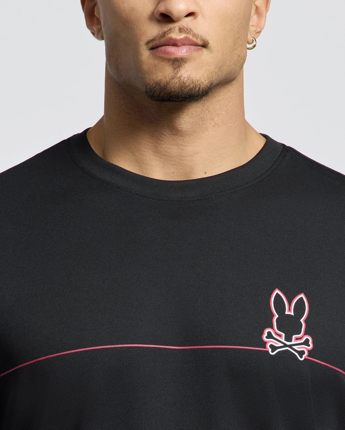 A person wears the Psycho Bunny MENS DACANO CHEST GRAPHIC SPORT TEE (B6U313E200) featuring a black color with a pink-outlined rabbit and crossbones logo, moisture-wicking technology, and a pink horizontal chest stripe. Short-haired, they complete their look with an earring.