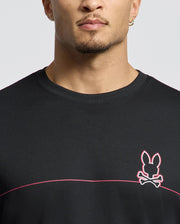 A person wears the Psycho Bunny MENS DACANO CHEST GRAPHIC SPORT TEE (B6U313E200) featuring a black color with a pink-outlined rabbit and crossbones logo, moisture-wicking technology, and a pink horizontal chest stripe. Short-haired, they complete their look with an earring.