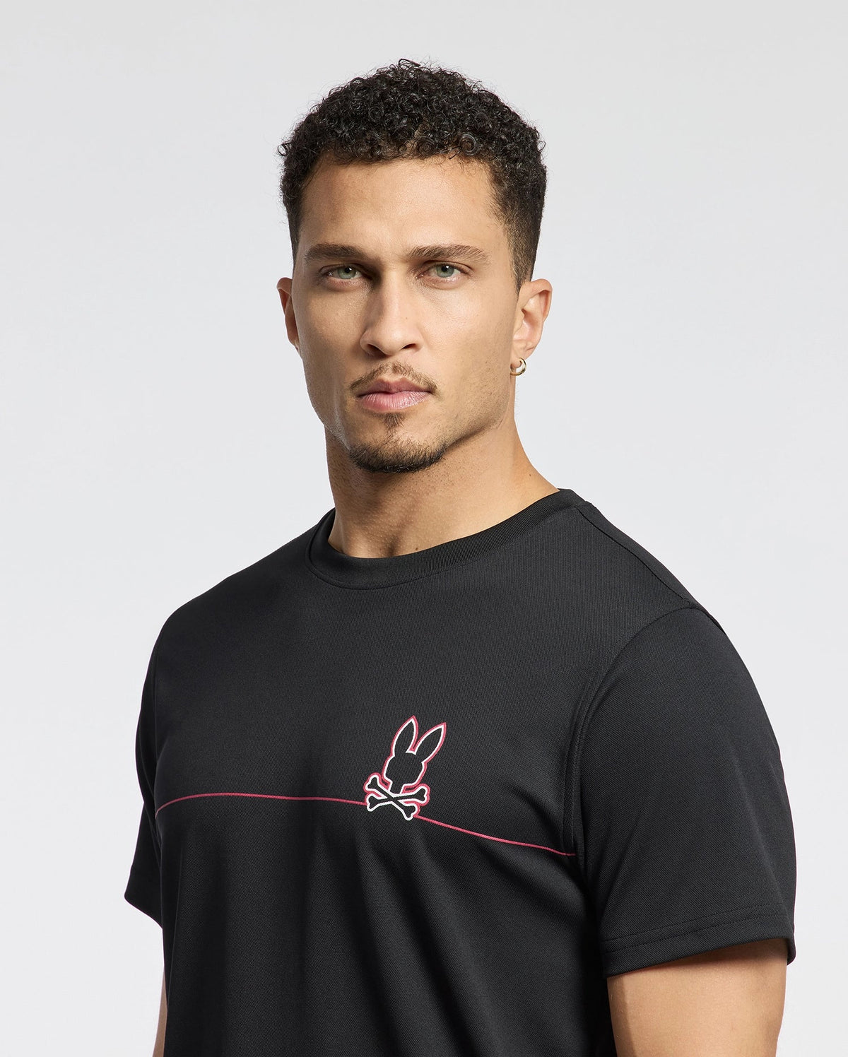 A man with short curly hair wears the Psycho Bunny MENS DACANO CHEST GRAPHIC SPORT TEE - B6U313E200. The black shirt features a pink rabbit and crossed bones emblem. Made from performance fabric with moisture-wicking technology, it ensures comfort during workouts against a plain white backdrop.