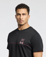 A man with short curly hair wears the Psycho Bunny MENS DACANO CHEST GRAPHIC SPORT TEE - B6U313E200. The black shirt features a pink rabbit and crossed bones emblem. Made from performance fabric with moisture-wicking technology, it ensures comfort during workouts against a plain white backdrop.