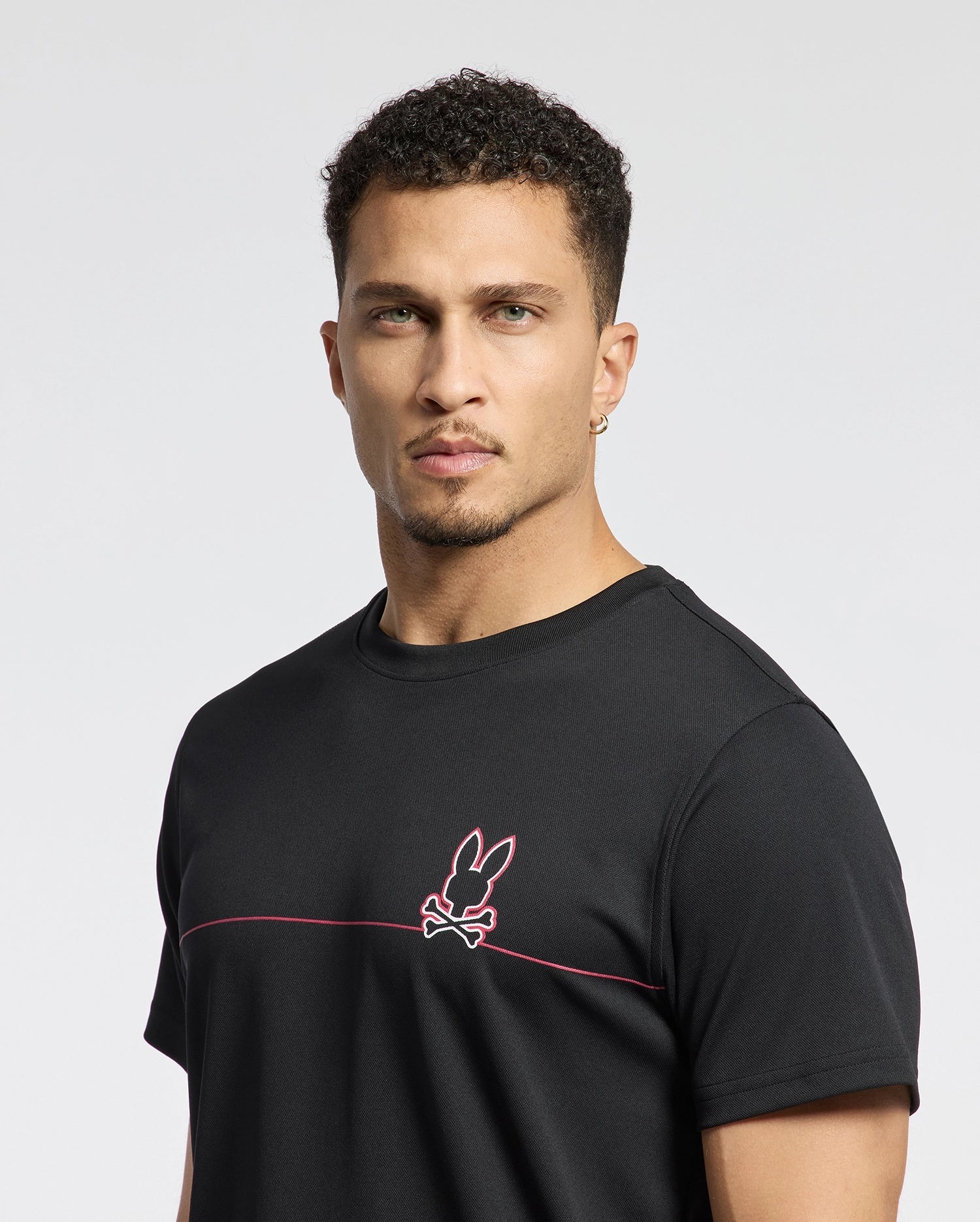 A man with short curly hair wears the Psycho Bunny MENS DACANO CHEST GRAPHIC SPORT TEE - B6U313E200. The black shirt features a pink rabbit and crossed bones emblem. Made from performance fabric with moisture-wicking technology, it ensures comfort during workouts against a plain white backdrop.