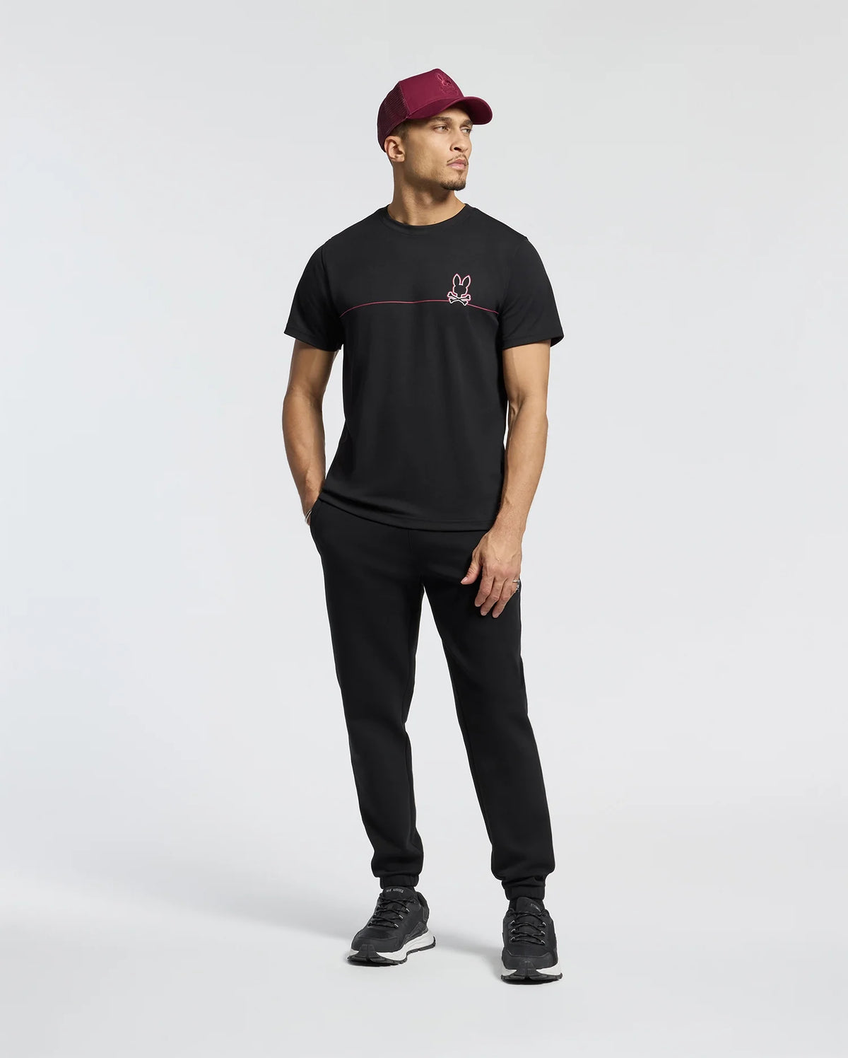 A man in a Psycho Bunny Men's Dacano Chest Graphic Sport Tee, black pants, moisture-wicking sneakers, and a maroon baseball cap stands in a relaxed pose against a light gray background.
