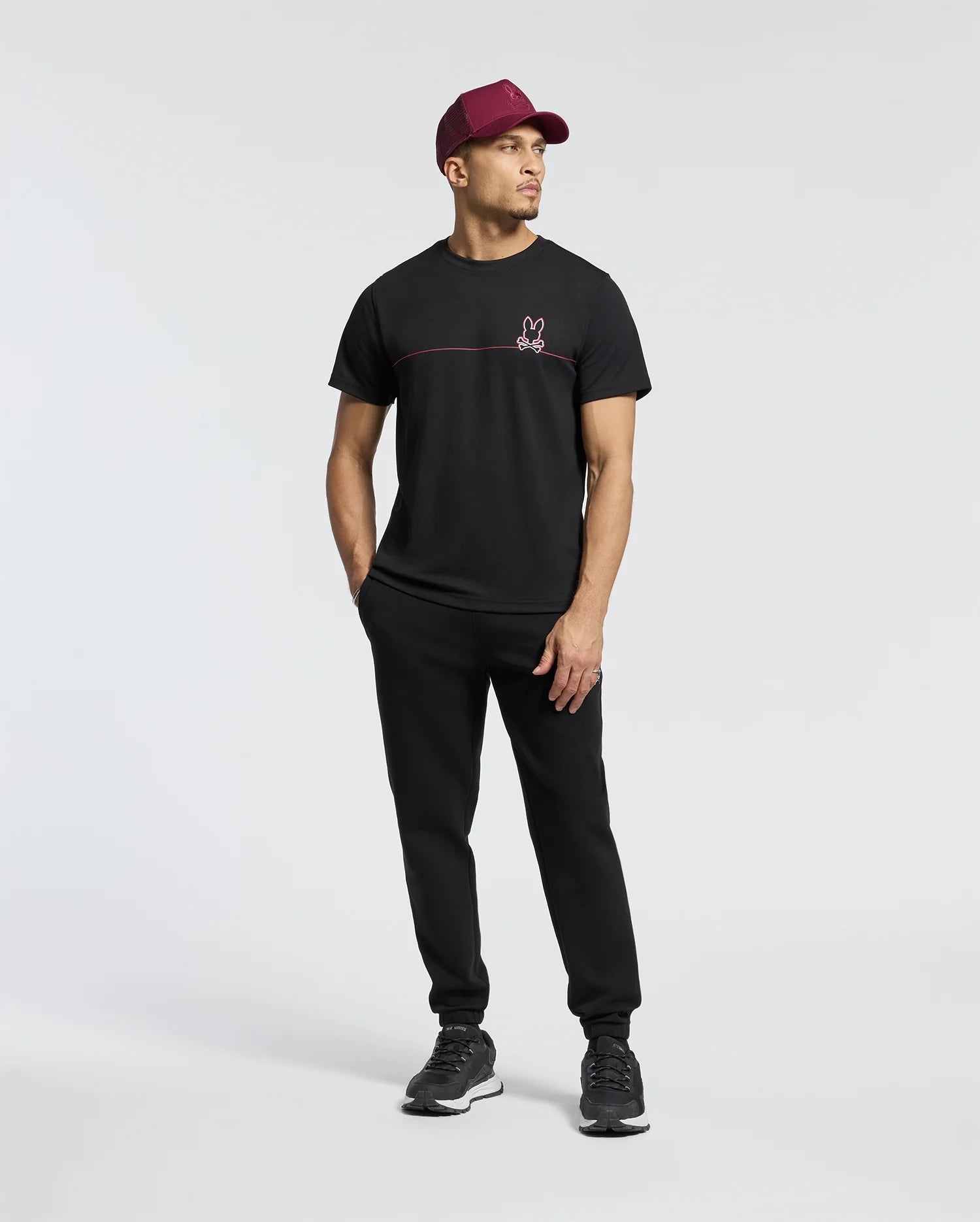 A man in a Psycho Bunny Men's Dacano Chest Graphic Sport Tee, black pants, moisture-wicking sneakers, and a maroon baseball cap stands in a relaxed pose against a light gray background.