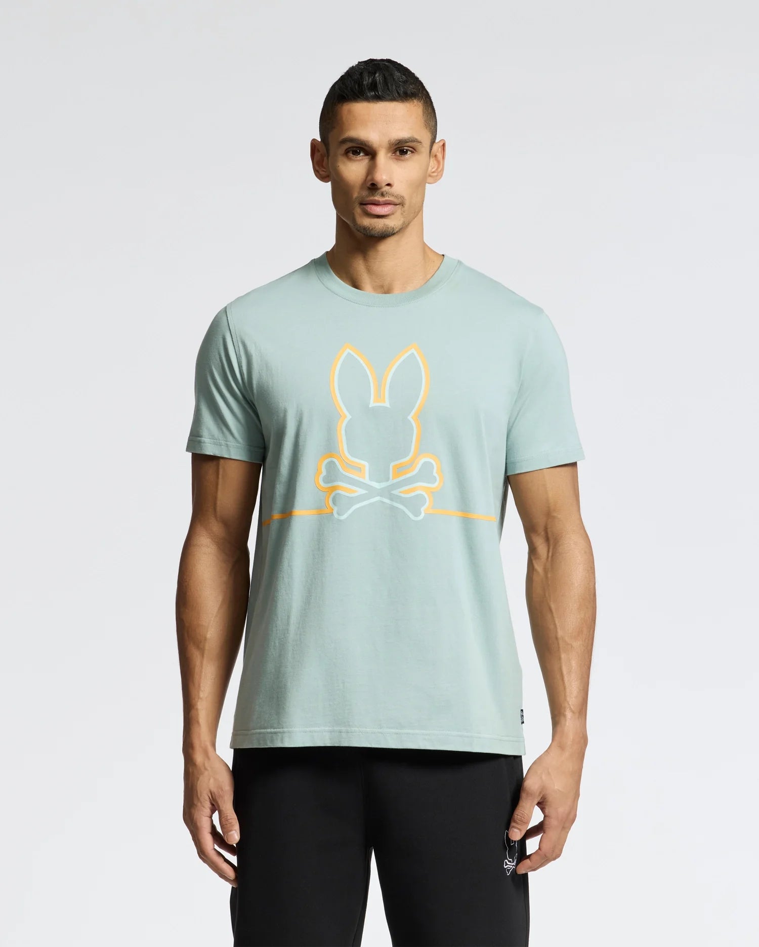 A man wears the Psycho Bunny MENS DACANO FRONT GRAPHIC TEE (B6U314E200) featuring a bunny and crossbones emblem. Its light green fabric, regular fit, and pairing with black pants create a comfortable style against a plain white background. Perfect for casual outings.
