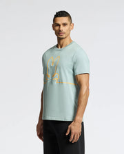 A person wearing Psycho Bunny's MENS DACANO FRONT GRAPHIC TEE - B6U314E200, a comfort-forward light blue T-shirt with an orange rabbit and crossbones design, pairs it with black pants against a plain white background. The regular fit Tee accentuates their neutral expression as they pose effortlessly.