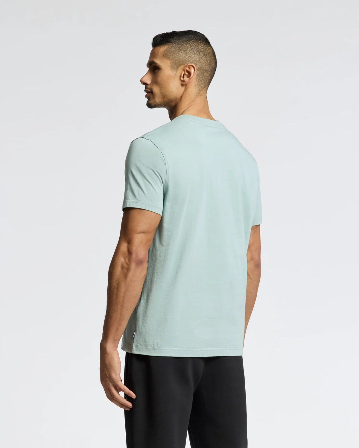 A man with short hair stands against a light gray background, looking to the side in Psycho Bunny's comfort-forward MENS DACANO FRONT GRAPHIC TEE - B6U314E200 in mint green and black pants. His arms rest relaxed at his sides.
