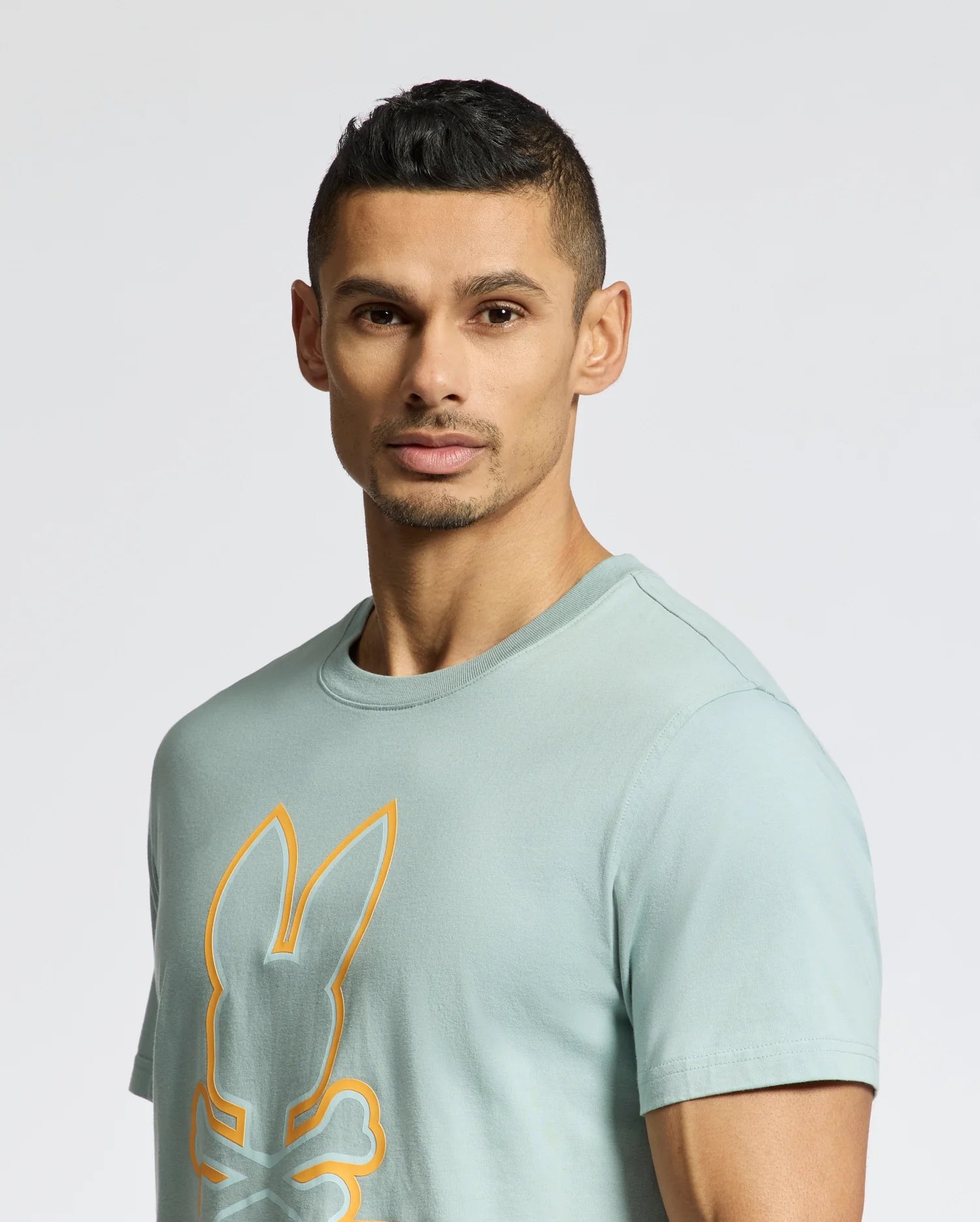 A person with short hair wears the light green MENS DACANO FRONT GRAPHIC TEE from Psycho Bunny, featuring an orange outline of a stylized bunny with crossed bones. The plain white background highlights the comfort-focused design of this unique piece.