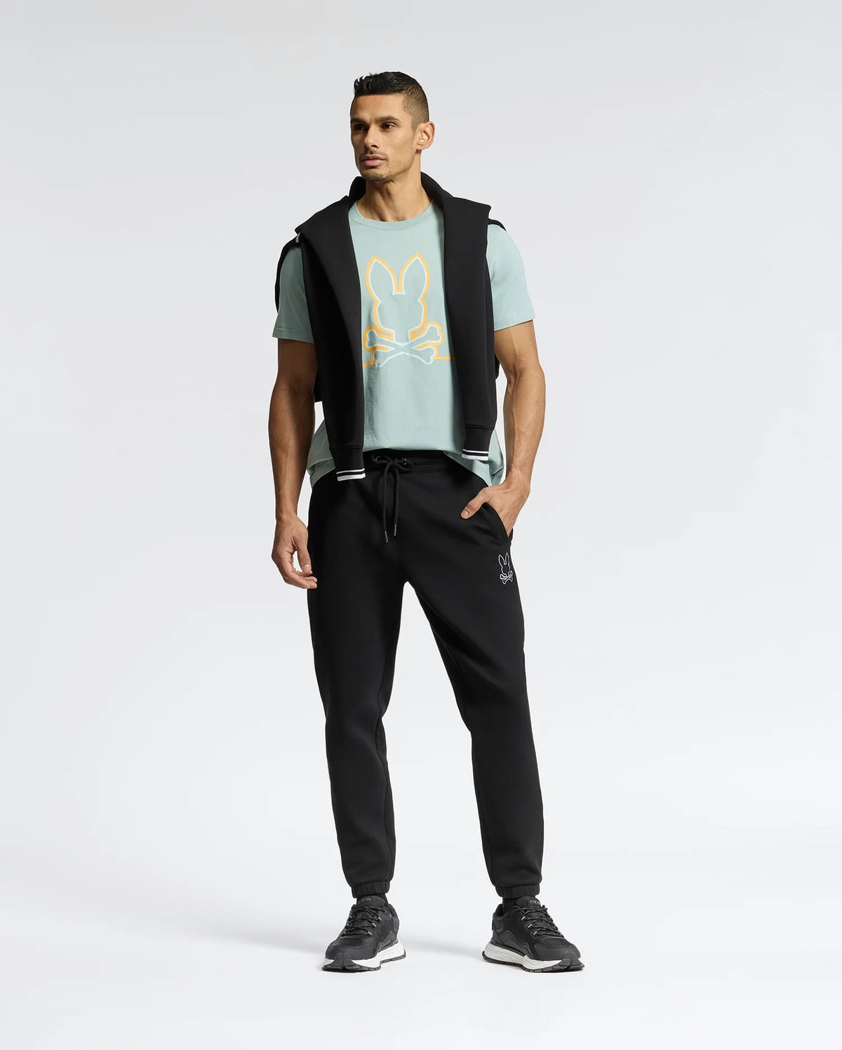 A man confidently stands in a Psycho Bunny MENS DACANO FRONT GRAPHIC TEE - B6U314E200, showcasing a stylized rabbit design. He's paired it with regular fit black jogger pants and sneakers, and has a black jacket draped over his shoulders against a minimalist background.