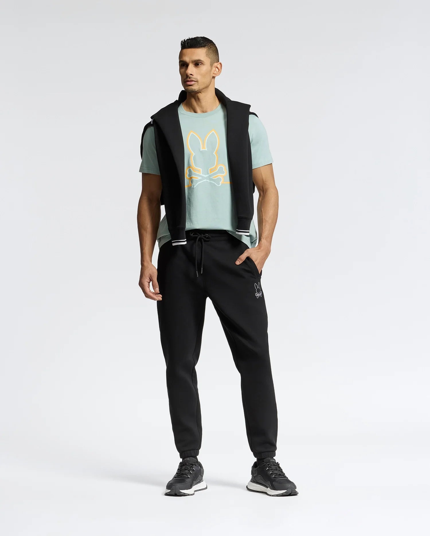 A man confidently stands in a Psycho Bunny MENS DACANO FRONT GRAPHIC TEE - B6U314E200, showcasing a stylized rabbit design. He's paired it with regular fit black jogger pants and sneakers, and has a black jacket draped over his shoulders against a minimalist background.