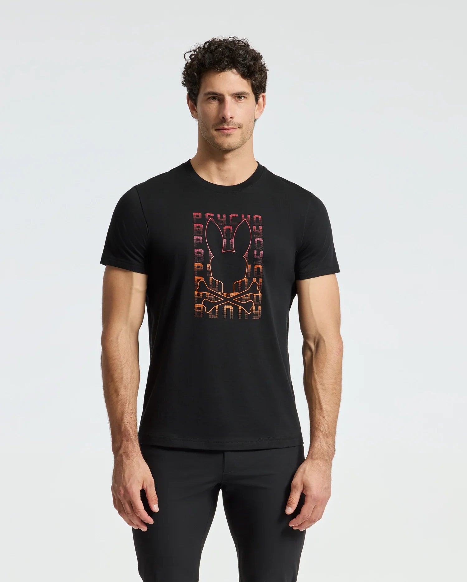 A man stands against a plain white background wearing the MENS CORTES GRAPHIC TEE - B6U328E200 by Psycho Bunny, which features a black T-shirt with an orange graphic of a skull and bunny ears.