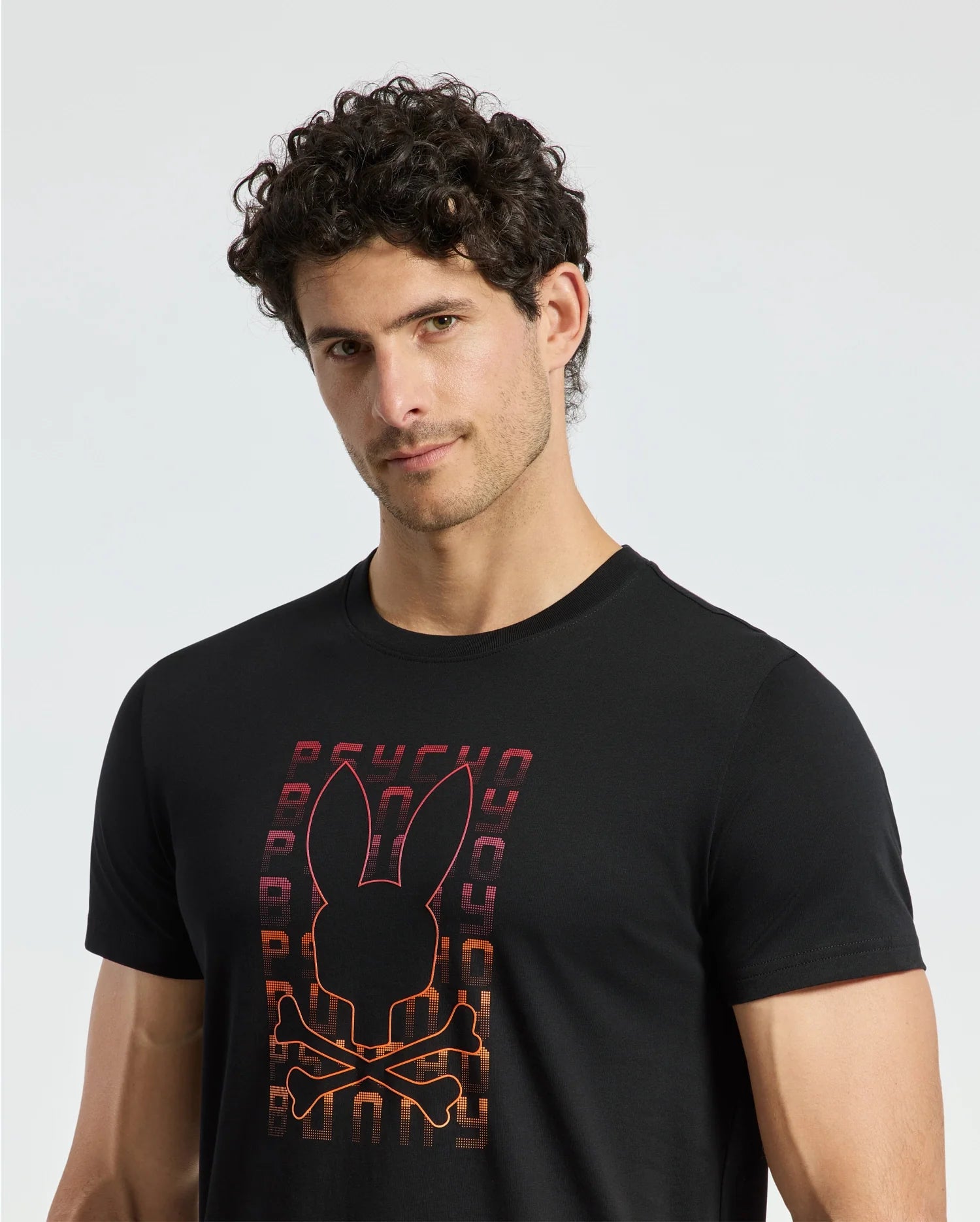A person with curly hair is wearing a MENS CORTES GRAPHIC TEE - B6U328E200 by Psycho Bunny. This lightweight cotton T-shirt showcases a bunny graphic with crossbones and stylized text, all set against a light, neutral background.
