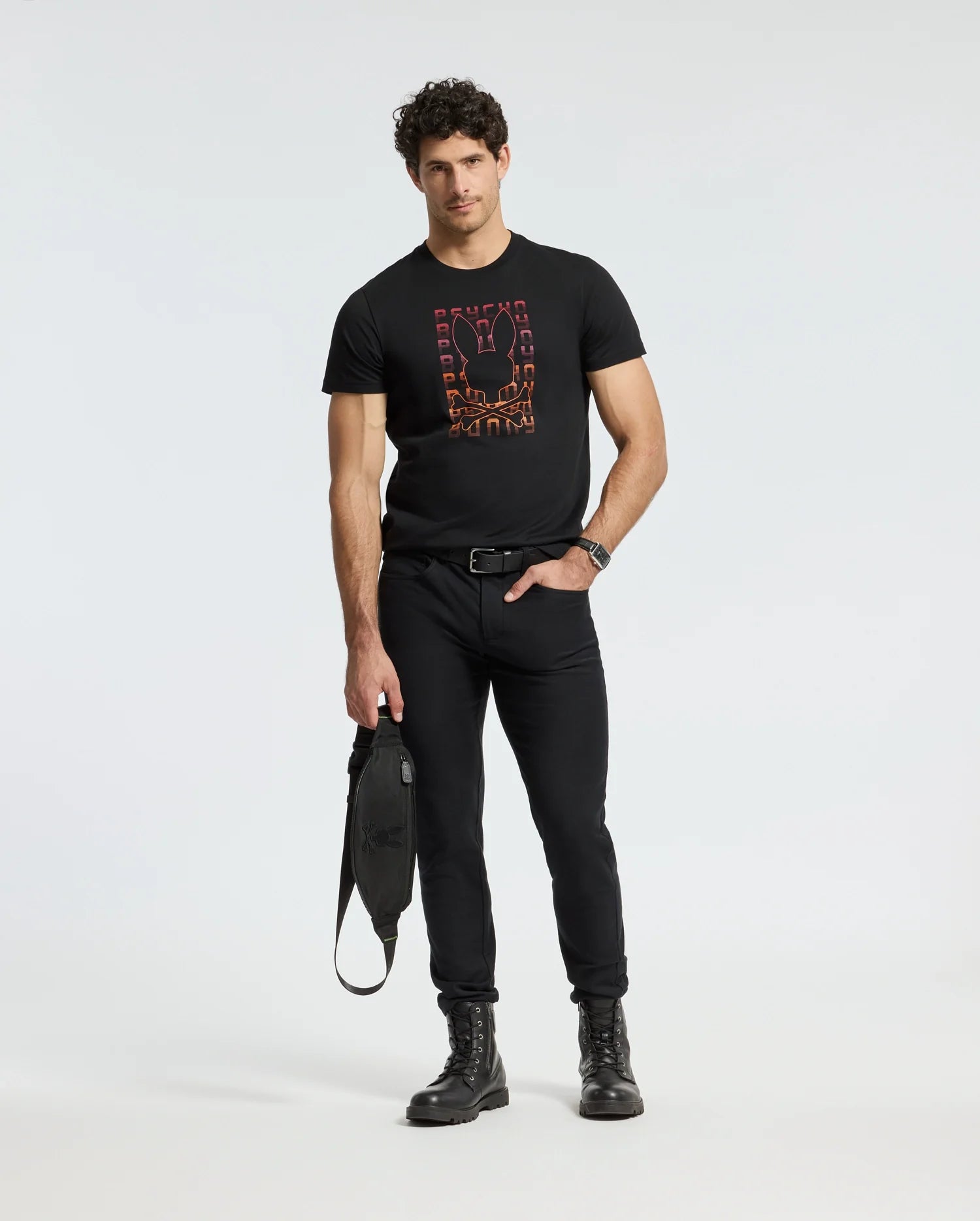 A man with curly hair stands confidently in a Psycho Bunny's MENS CORTES GRAPHIC TEE - B6U328E200, paired with black jeans and boots, holding a sleek black bag. He looks directly at the camera against a plain white background.
