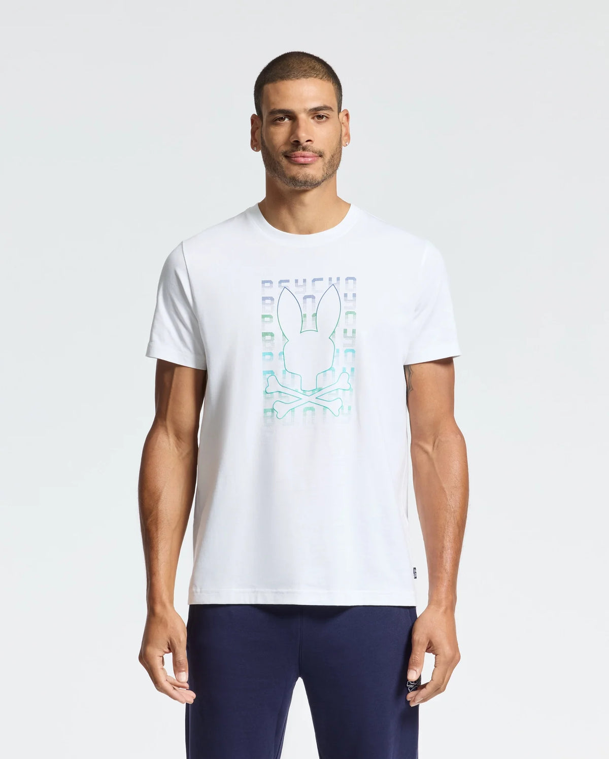A man wearing Psycho Bunny's Men's Cortes Graphic Tee - B6U328E200, featuring a rabbit and geometric design on the front, pairs it with dark pants while standing against a plain background.