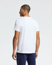 A man with short hair stands facing away, wearing the Psycho Bunny MENS CORTES GRAPHIC TEE - B6U328E200 with a minimalist design. Paired with dark blue pants, the outfit contrasts subtly against a light gray background.