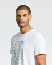 A man with short hair and an earring models the Psycho Bunny MENS CORTES GRAPHIC TEE - B6U328E200 featuring a bunny head and crossbones. Against a plain light background, he gazes confidently at the camera, highlighting his style.