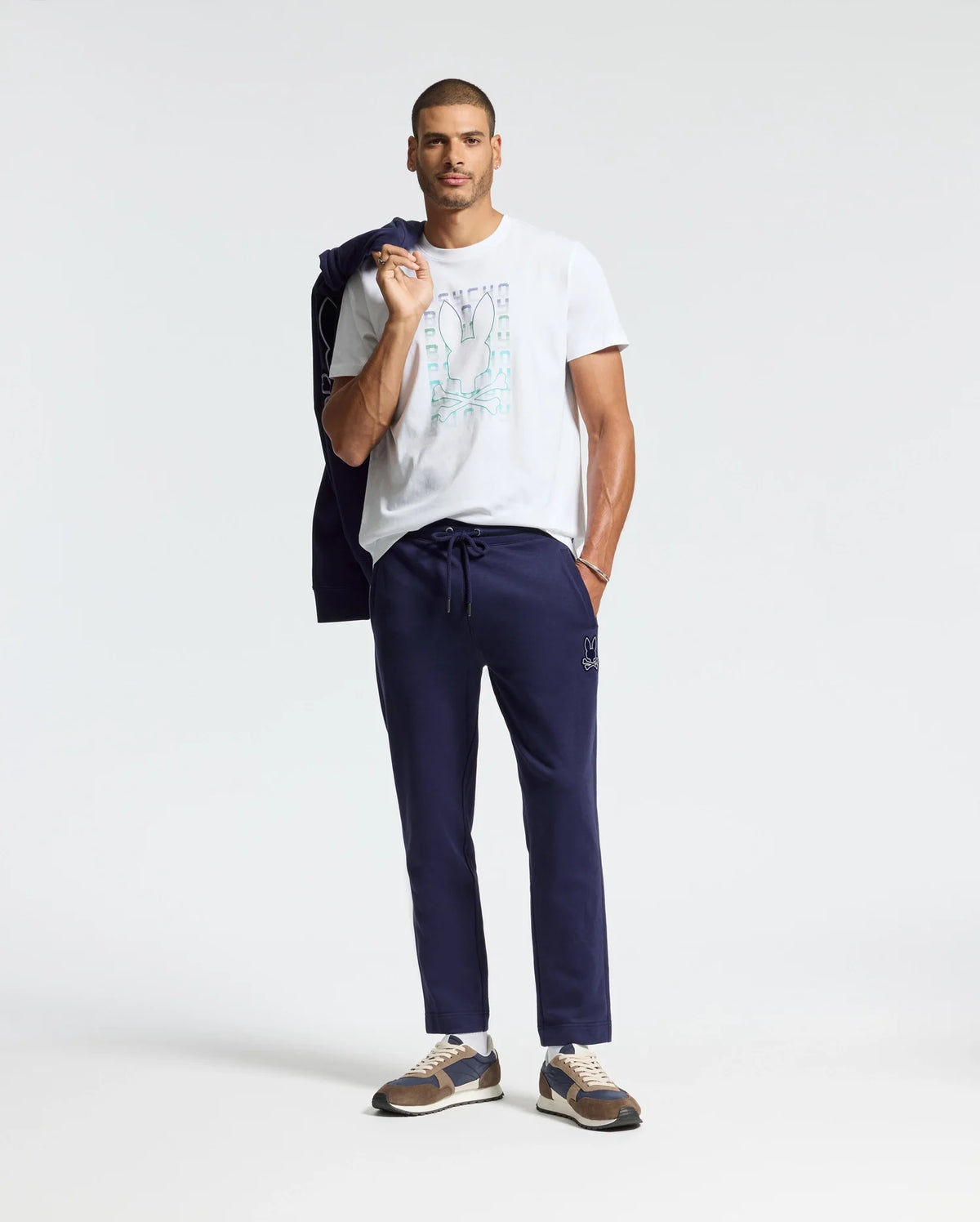 A man casually poses in a white Psycho Bunny Men's Cortes Graphic Tee, paired with navy jogger pants and sneakers. He drapes a navy jacket over his shoulder, and his short hair adds to the laid-back vibe against the plain white background, highlighting his effortlessly cool style.