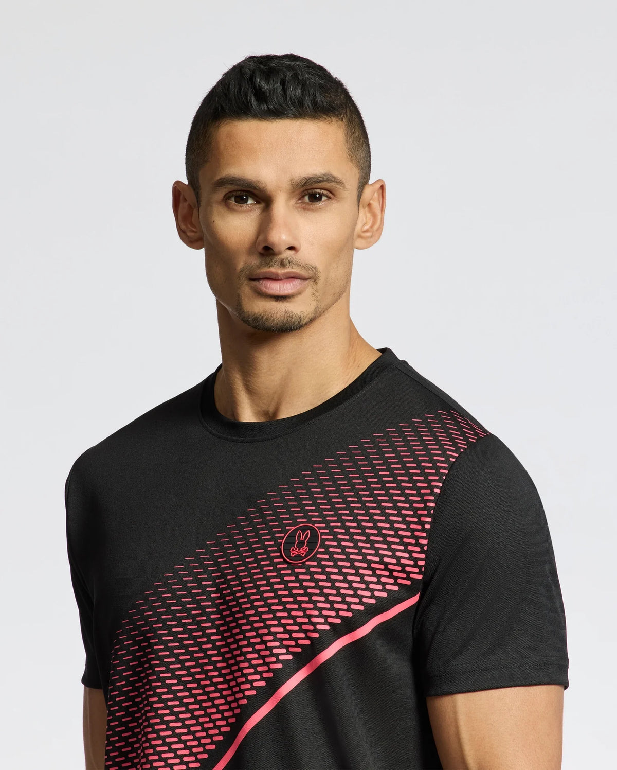 A person wearing a black Psycho Bunny MENS MACK SPORT TEE (B6U335E200) with a red geometric chest pattern stands against a plain background. The Moisture Wicking fabric complements their short, dark hair and neutral expression, highlighting both comfort and style.