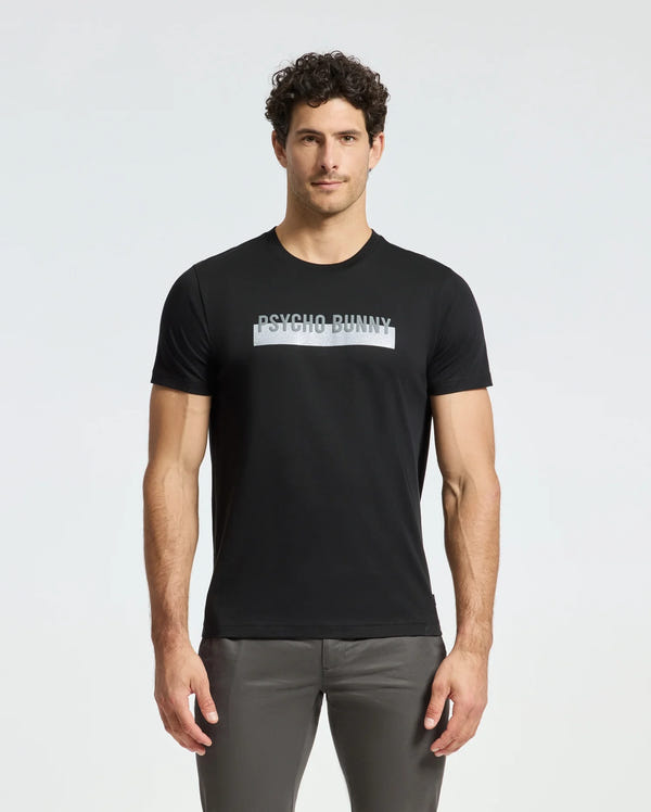 A man stands against a plain white background, wearing the MENS BROOKPORT GRAPHIC TEE - B6U362E200 by Psycho Bunny. This black graphic tee, crafted from premium quality Pima cotton, prominently features the "Psycho Bunny" design on the front, offering a stylish and comfortable modern look.