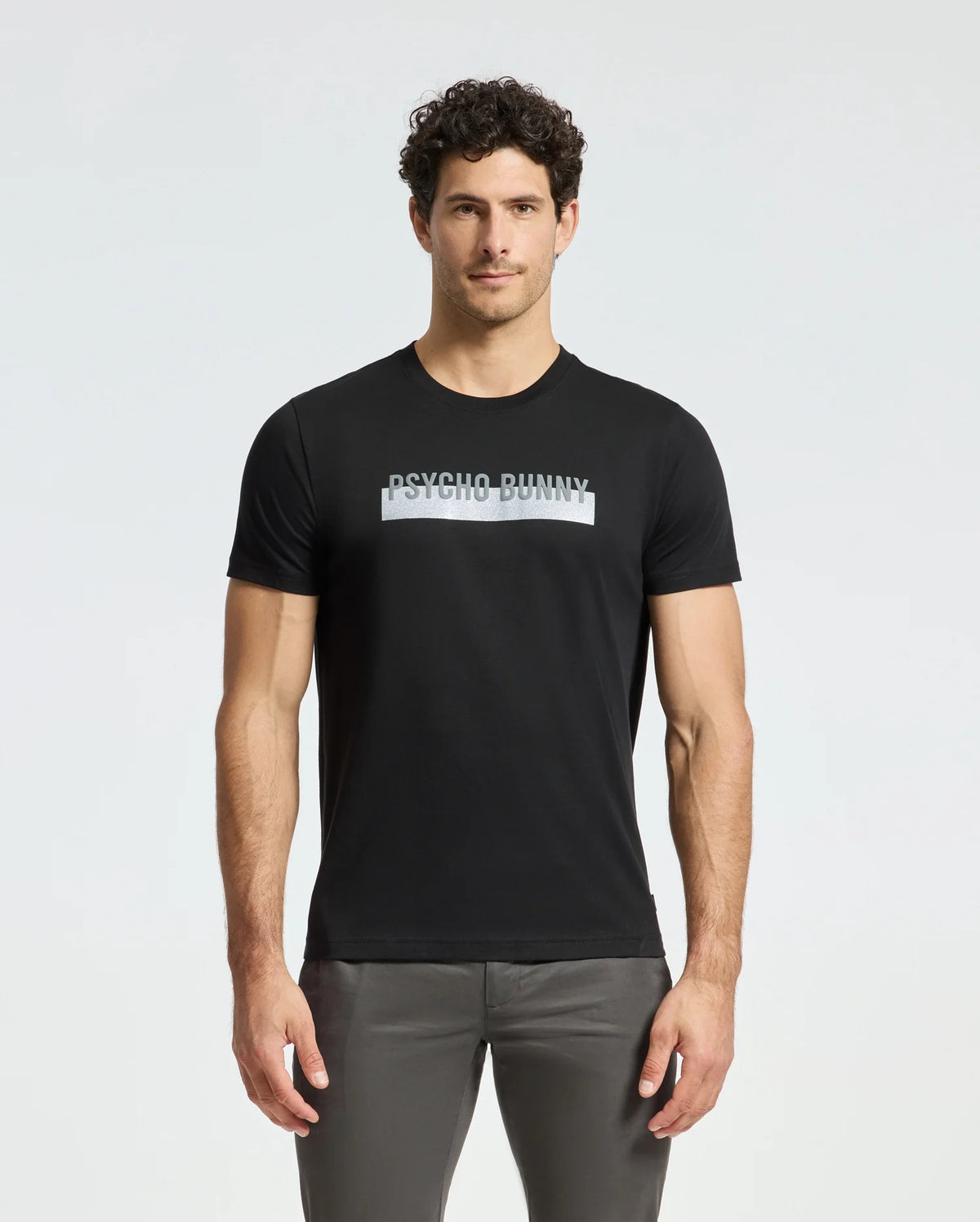A man stands against a plain white background, wearing the MENS BROOKPORT GRAPHIC TEE - B6U362E200 by Psycho Bunny. This black graphic tee, crafted from premium quality Pima cotton, prominently features the "Psycho Bunny" design on the front, offering a stylish and comfortable modern look.
