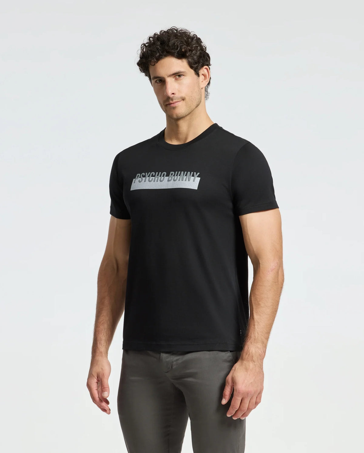 A man with curly hair is sporting the Psycho Bunny Men's Brookport Graphic Tee (B6U362E200) in black, made from premium Pima cotton, and has paired it stylishly with gray pants against a plain white background.