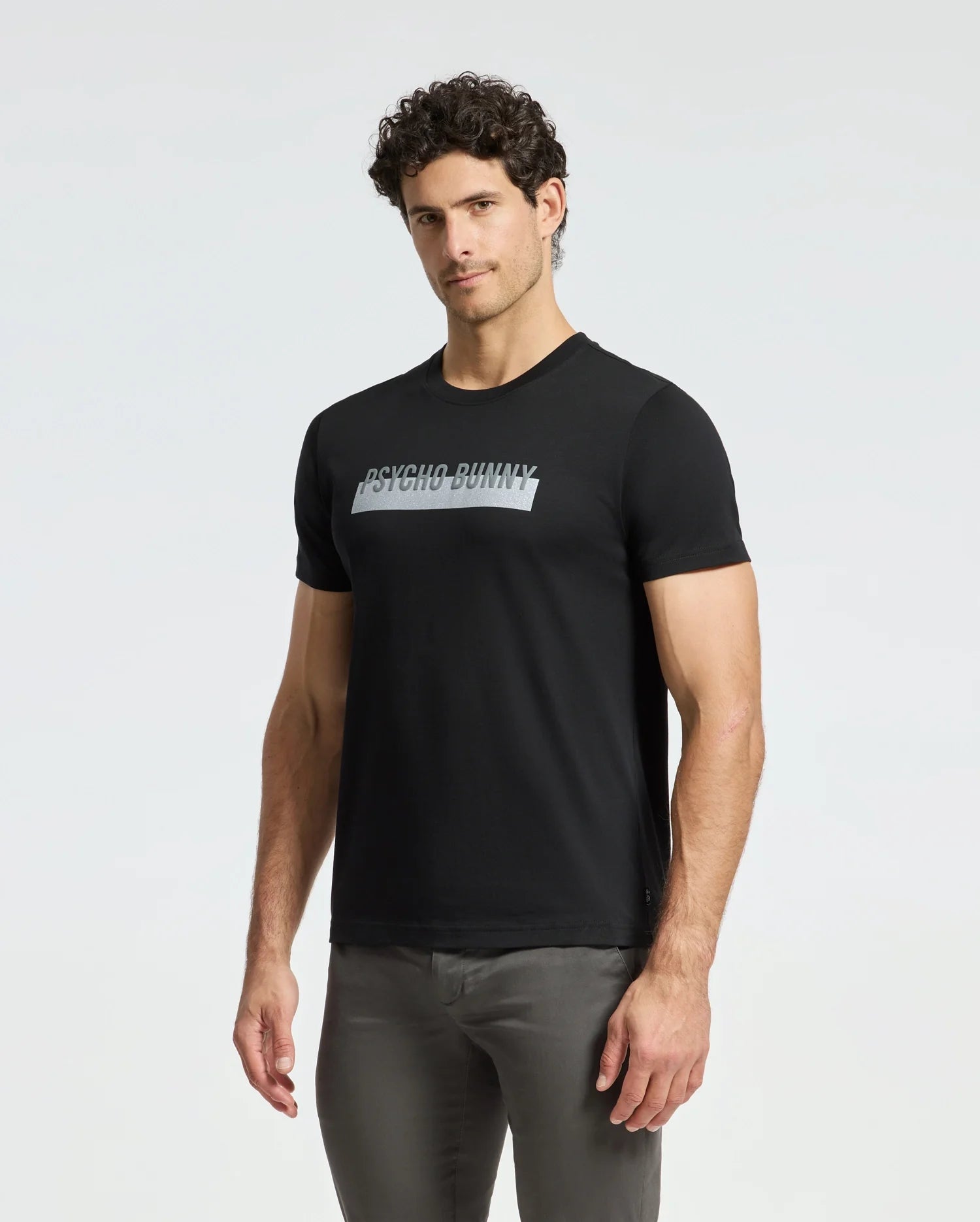 A man with curly hair is sporting the Psycho Bunny Men's Brookport Graphic Tee (B6U362E200) in black, made from premium Pima cotton, and has paired it stylishly with gray pants against a plain white background.