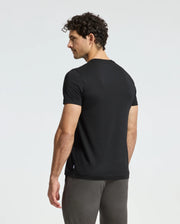 A man with curly hair stands facing slightly away from the camera, wearing a Psycho Bunny MENS BROOKPORT GRAPHIC TEE - B6U362E200, crafted from premium quality Pima cotton, paired with dark pants against a plain background.