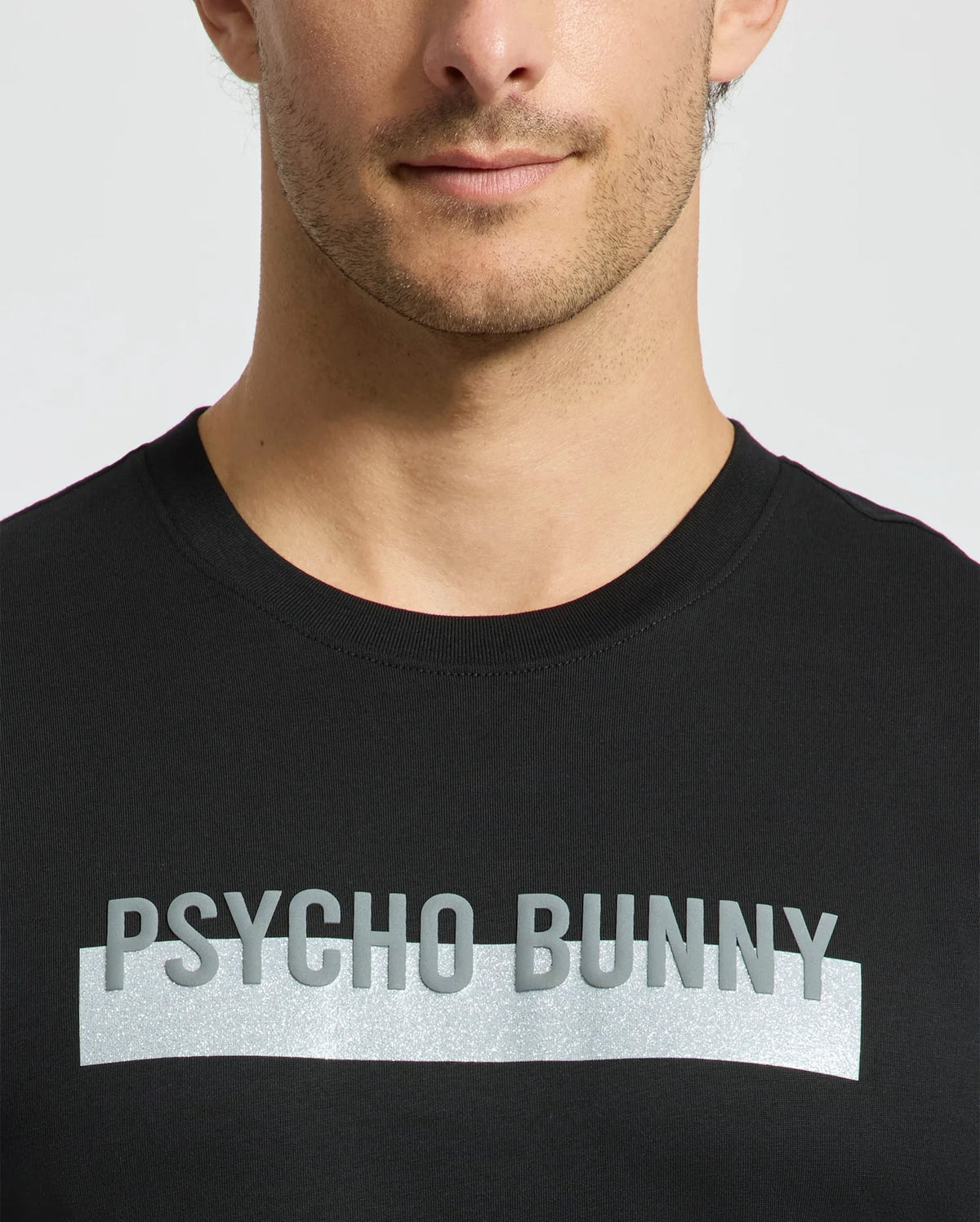 A man wearing the men's black Brookport Graphic Tee by Psycho Bunny, featuring "PSYCHO BUNNY" in silver lettering across the chest, stands out against the plain background.