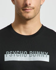 A man wearing the men's black Brookport Graphic Tee by Psycho Bunny, featuring "PSYCHO BUNNY" in silver lettering across the chest, stands out against the plain background.