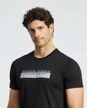 A person with curly hair stands against a plain backdrop wearing a Men's Brookport Graphic Tee by Psycho Bunny, in black, with "Psycho Bunny" printed on the front. This premium quality T-shirt, identified as B6U362E200, is crafted from soft Pima Cotton and adds an edge to any casual look.