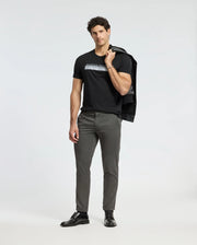 A man stands confidently with a jacket slung over his shoulder. He sports the Psycho Bunny MENS BROOKPORT GRAPHIC TEE in black, complemented by gray pants and black shoes. The plain white background highlights his casual yet stylish outfit, emphasizing the premium quality of his attire.