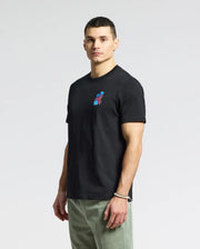 A man stands against a plain backdrop wearing the MENS PUERTO RICO TEE - B6U382F200 by Psycho Bunny, a black Pima cotton t-shirt with a colorful chest graphic showing his Puerto Rican pride. Olive green pants and a bracelet on his right wrist complete the look as he gazes neutrally at the camera.