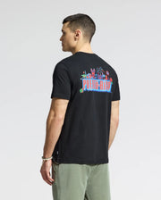 A person in a Psycho Bunny MENS PUERTO RICO TEE - B6U382F200, featuring soft Pima cotton and vibrant "Puerto Rico" graphics on the back, pairs it with olive green pants. Standing against a plain background, they exude Puerto Rican pride while glancing to the side.