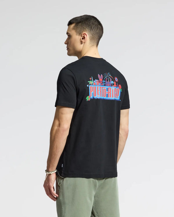 A person in a Psycho Bunny MENS PUERTO RICO TEE - B6U382F200, featuring soft Pima cotton and vibrant "Puerto Rico" graphics on the back, pairs it with olive green pants. Standing against a plain background, they exude Puerto Rican pride while glancing to the side.
