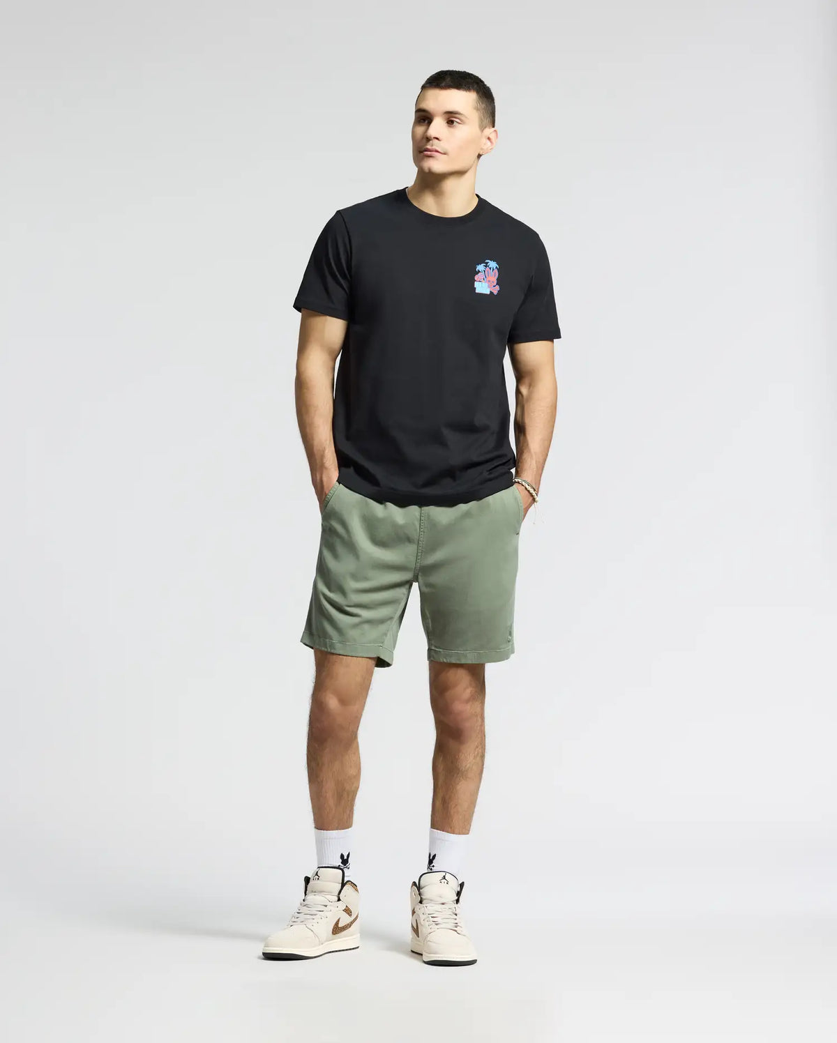 Against a blank backdrop, an individual wears a black Psycho Bunny MENS PUERTO RICO TEE - B6U382F200 with a vibrant Puerto Rican-inspired chest design, paired with green shorts, white socks, and beige sneakers. Their hands rest in their pockets, and they maintain a neutral expression.