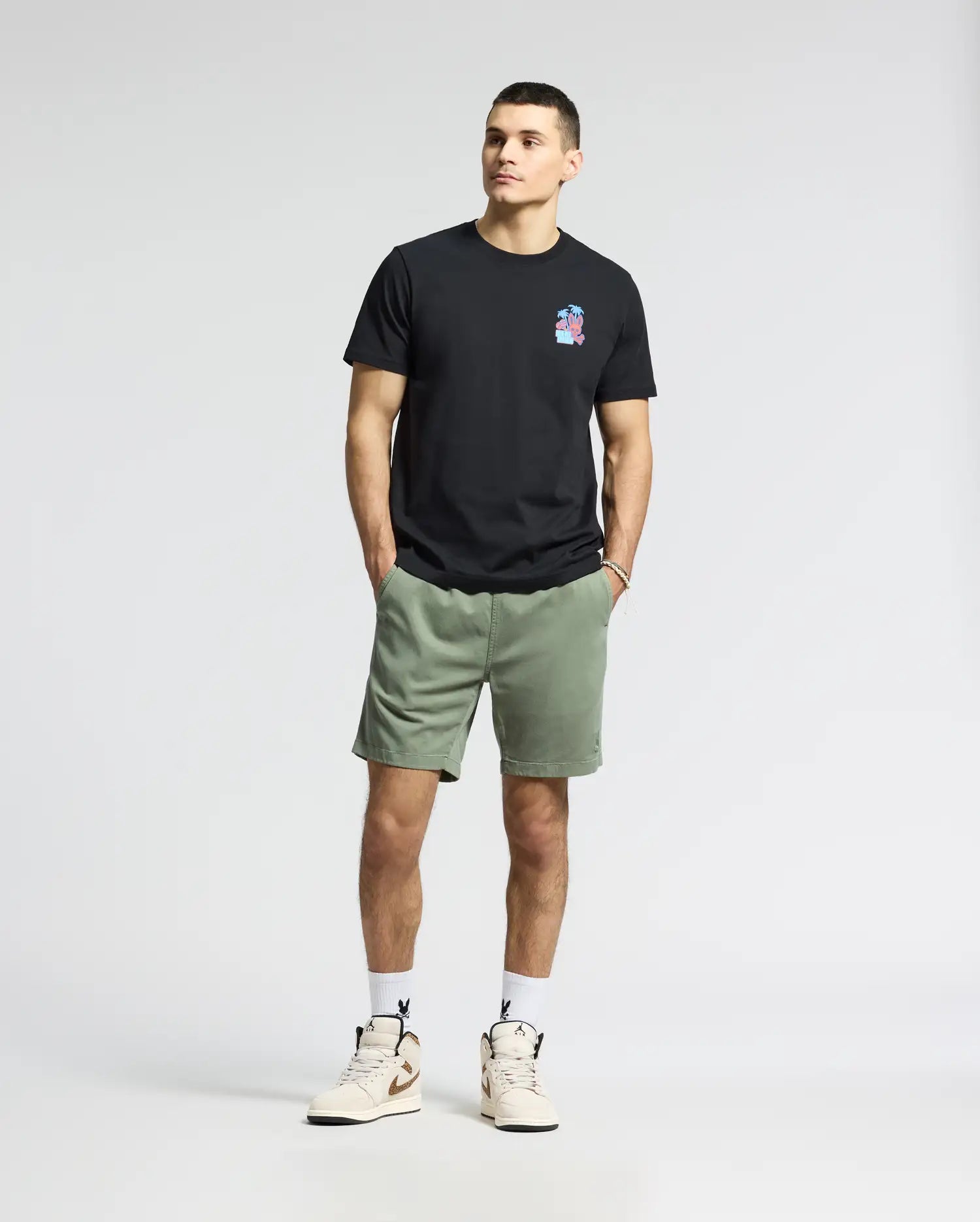 Against a blank backdrop, an individual wears a black Psycho Bunny MENS PUERTO RICO TEE - B6U382F200 with a vibrant Puerto Rican-inspired chest design, paired with green shorts, white socks, and beige sneakers. Their hands rest in their pockets, and they maintain a neutral expression.