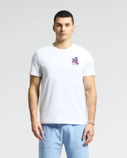 A man in a white Psycho Bunny MENS PUERTO RICO TEE - B6U382F200 featuring a small beach graphic on the chest, made from soft Pima cotton, is paired with light blue pants and stands against a plain background.