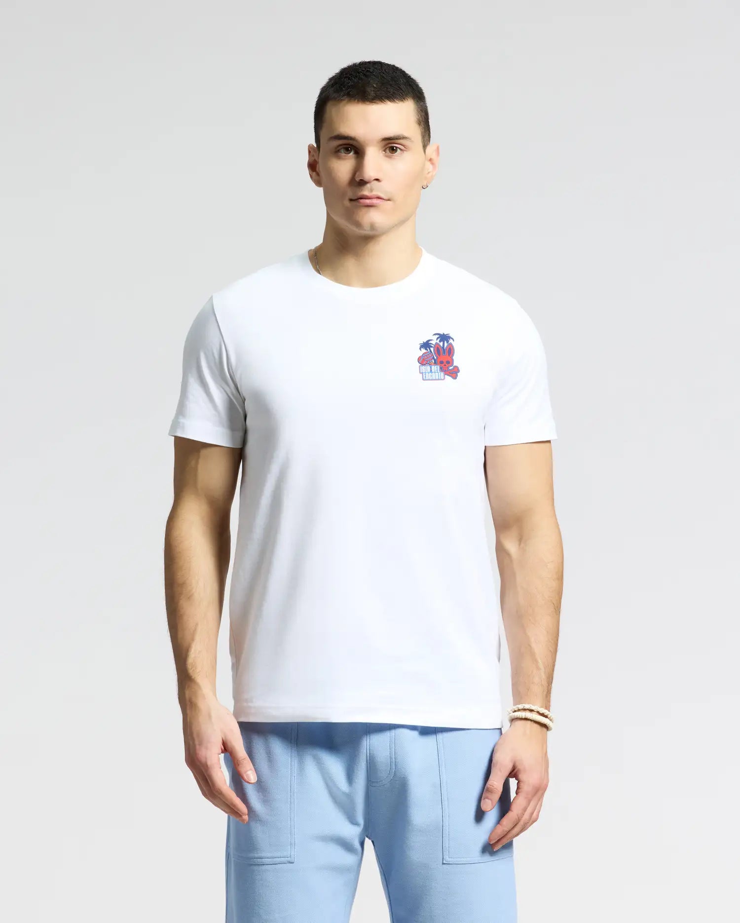 A man in a white Psycho Bunny MENS PUERTO RICO TEE - B6U382F200 featuring a small beach graphic on the chest, made from soft Pima cotton, is paired with light blue pants and stands against a plain background.